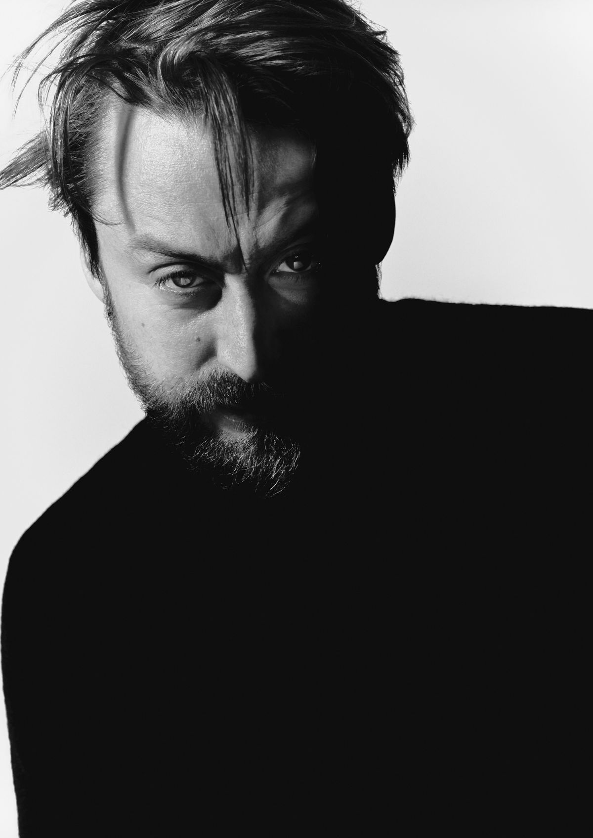 Monochrome portrait of a bearded man with intense gaze, partially lit, wearing a dark sweater against a white background.