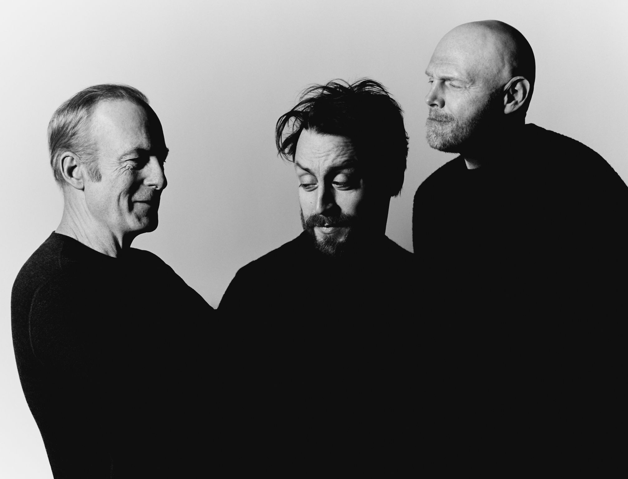 Black and white studio portrait featuring three adult males in dark sweaters against a plain background, looking contemplative and relaxed.