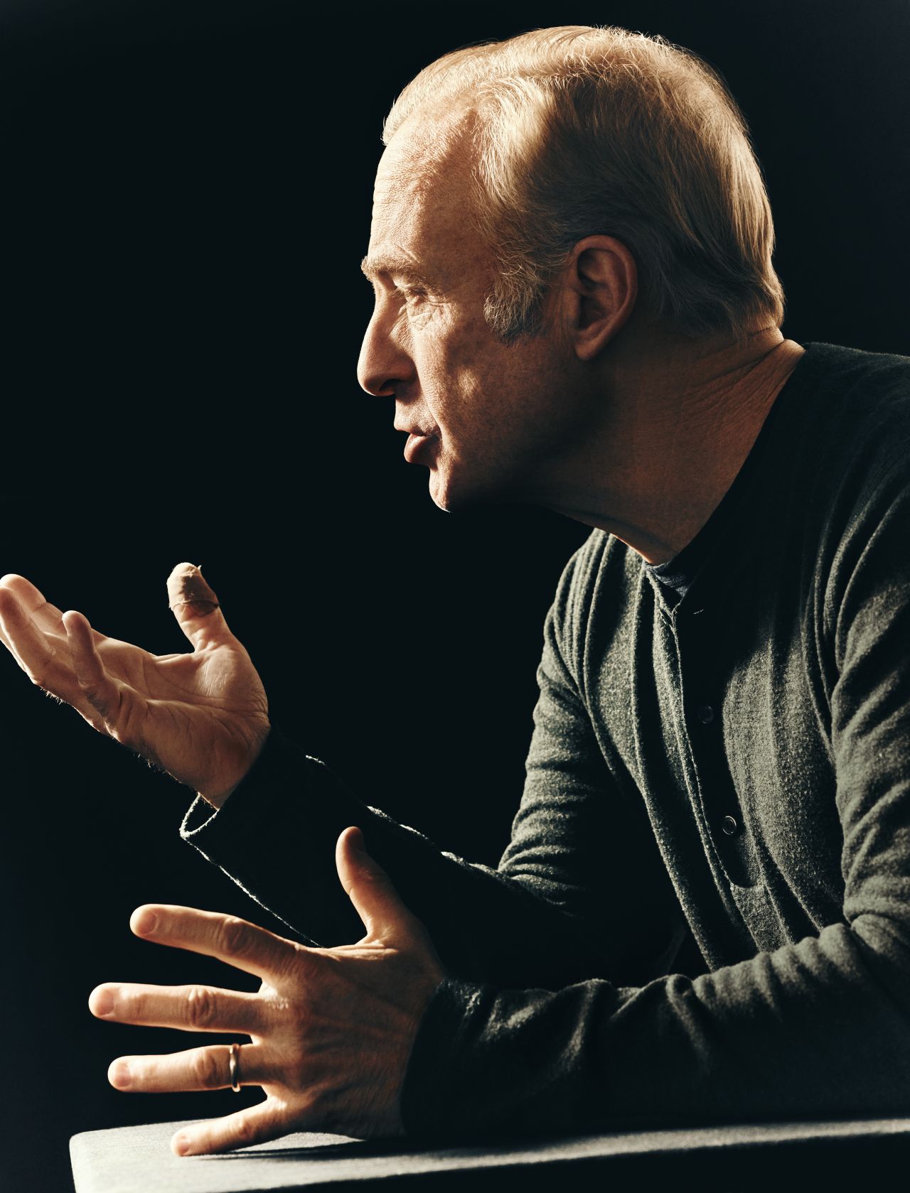 Profile shot of an adult male in a dark grey shirt speaking with his hands up, set against a dark background in a studio setting.