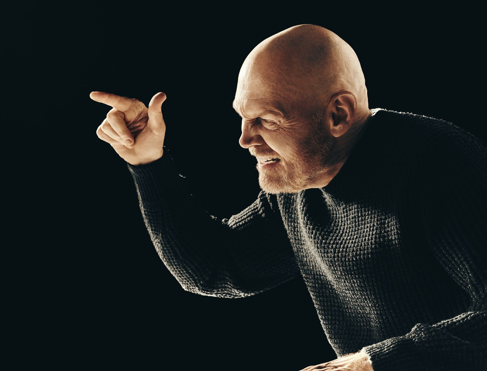 Middle-aged man with a bald head in a dark sweater, pointing with a stern expression against a dark background, creating a dramatic portrait.