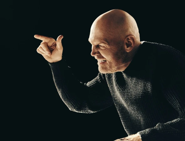 Middle-aged man with a bald head in a dark sweater, pointing with a stern expression against a dark background, creating a dramatic portrait.