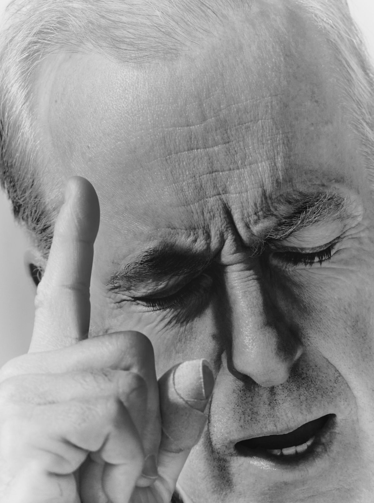 Monochromatic close-up captures a mature man with furrowed brow, eyes closed, finger raised as if deep in thought, against a white background.