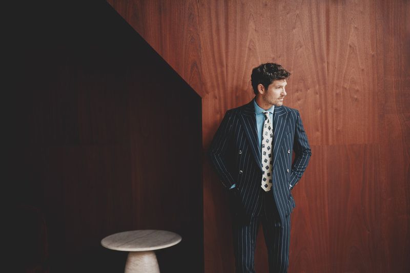 Man in a pinstripe suit stands indoors against a paneled wall. He wears a patterned tie and a light-blue shirt, styled in editorial fashion.
