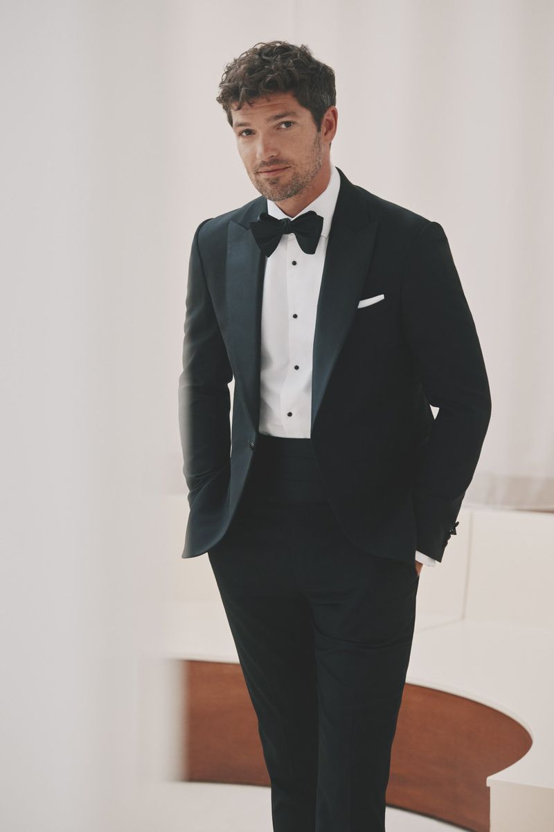 Portrait of a handsome adult male posing in a black tuxedo with a bow tie, standing in a well-lit indoor setting.