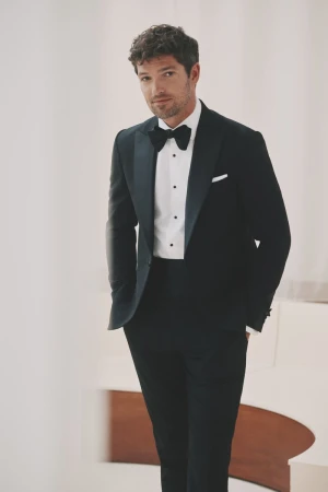 Portrait of a handsome adult male posing in a black tuxedo with a bow tie, standing in a well-lit indoor setting.