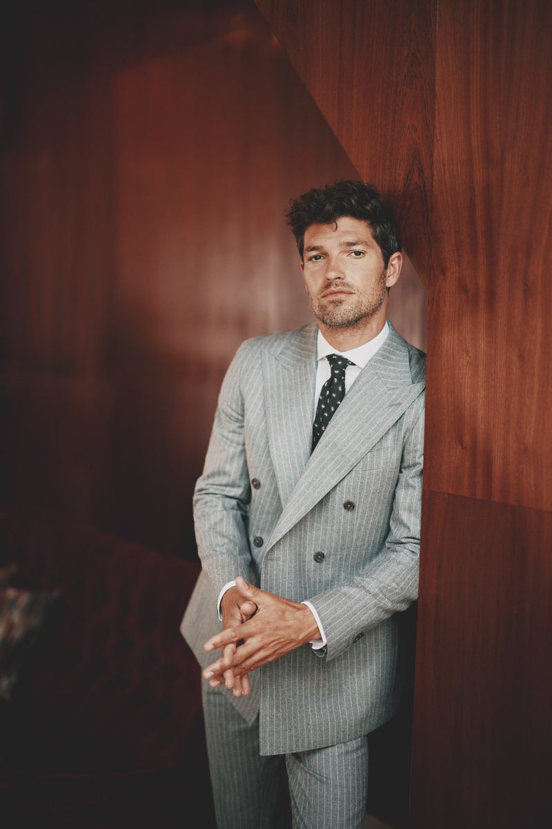 Portrait of a man in a pinstripe suit standing indoors; he's leaning against a wood wall while looking at the camera.