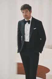 Man wearing a black tuxedo with a white dress shirt and black bowtie against a bright studio background