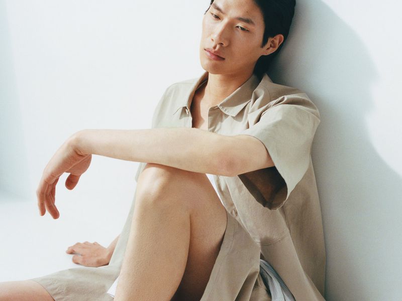 Portrait of a young Asian man in a beige short-sleeve shirt and shorts sitting on the floor against a white background.