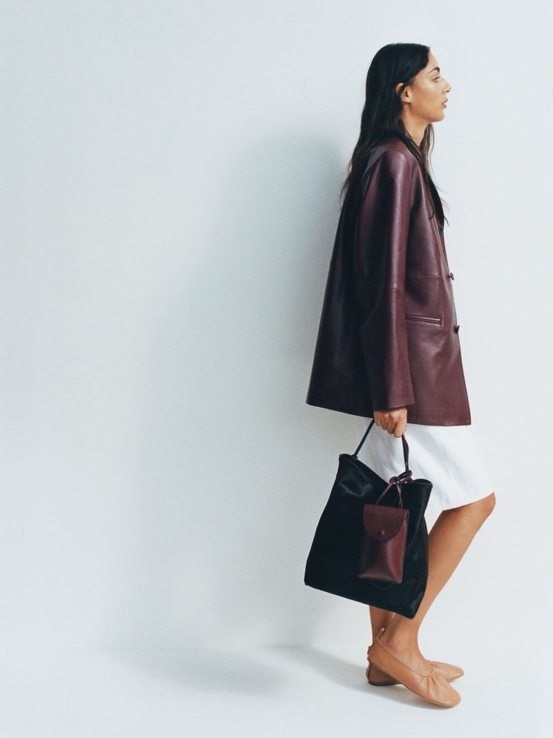 A woman models a burgundy jacket and white dress, carrying a dark tote bag against a seamless white backdrop in a studio setting.
