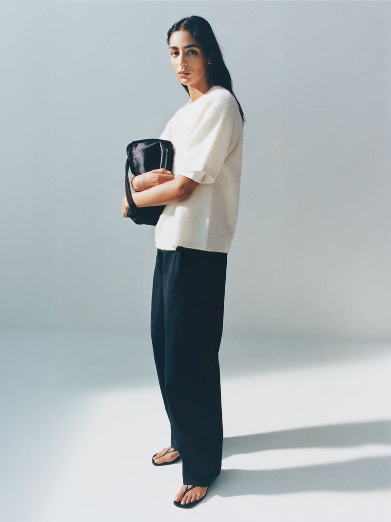 Adult female in a photo studio poses in a white top and black pants, holding a black bag in a fashion or beauty context.
