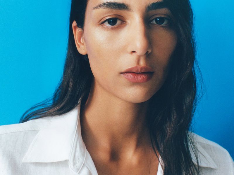 Close-up portrait of a young adult woman with dark hair wearing a white collared shirt against a bright blue background in studio lighting.