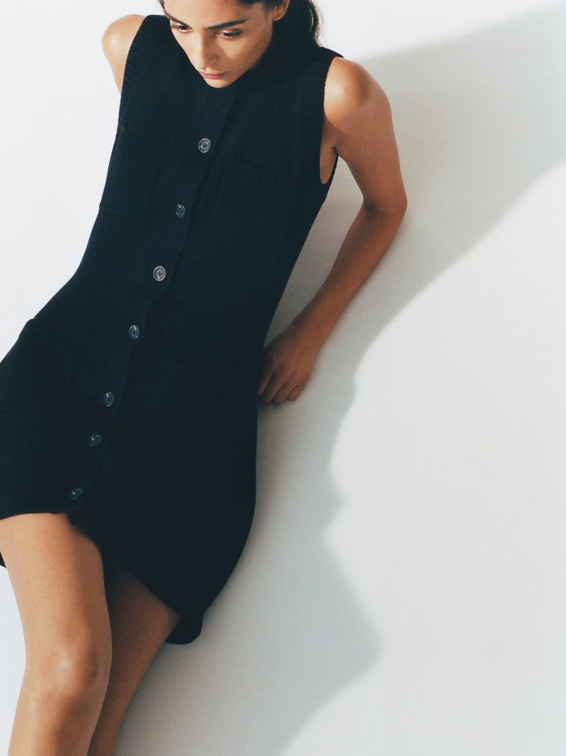 Woman wearing a sleeveless black dress with buttons, leaning against a white wall in a studio setting, creating a minimalist fashion shot.