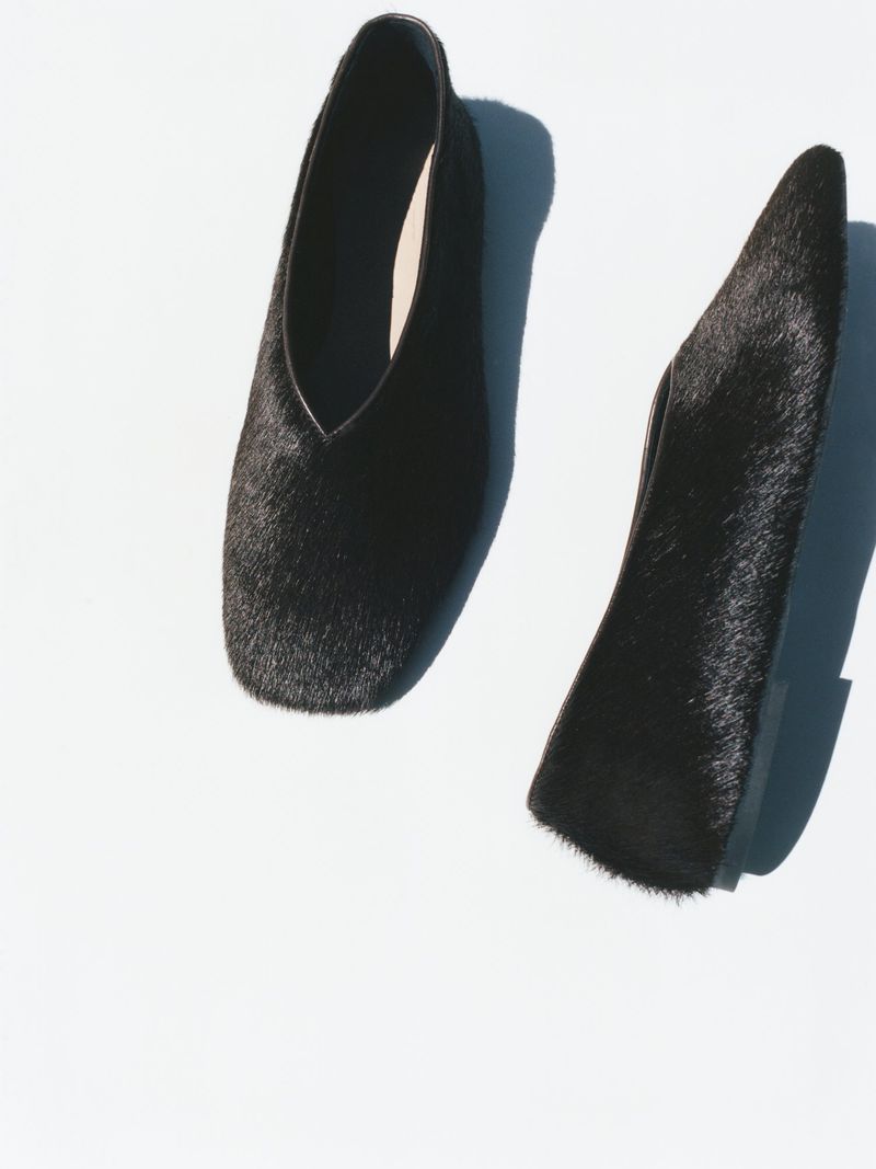 Close-up product shot showcases two black, fur-covered, pointed-toe heeled shoes on a white backdrop with natural shadows. Fashion.