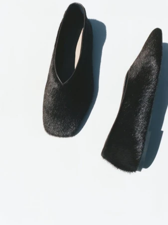 Close-up product shot showcases two black, fur-covered, pointed-toe heeled shoes on a white backdrop with natural shadows. Fashion.