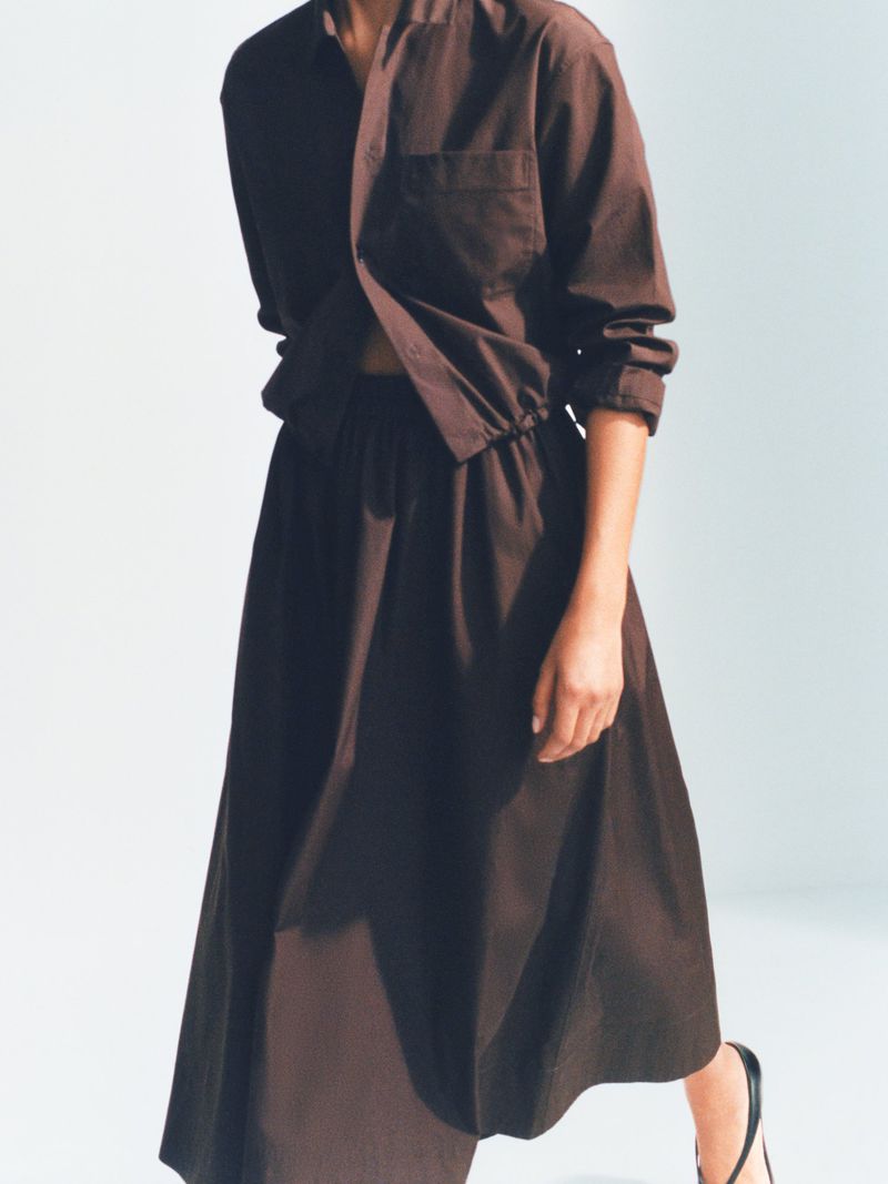 Woman posing in an oversized brown collared shirt and matching midi skirt, accessorized with black sandals, against a neutral backdrop.
