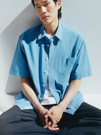 Stylish Asian man in a blue button-down shirt and dark pants sits indoors against a white wall, radiating casual fashion and comfort.