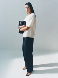 Model Nora Attal wearing a cream t-shirt and black trousers holding a dark bag against a white studio background