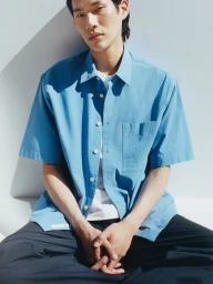 A man wearing a blue short-sleeve button-down shirt and dark trousers sitting against a white studio wall.