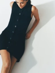 Model Nora Attal posing from an overhead angle in a black sleeveless button-up mini dress against a white studio background.