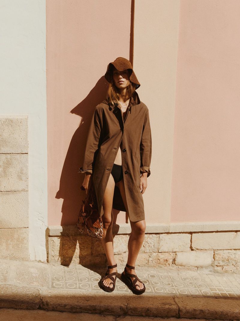 Model Lea Seelenmeyer wearing a brown coat, sun hat, and strappy sandals standing outdoors against a pink stucco wall.