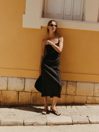Woman in a black dress and sunglasses stands against an ochre wall outdoors, holding a bag, in a fashion or lifestyle shot.