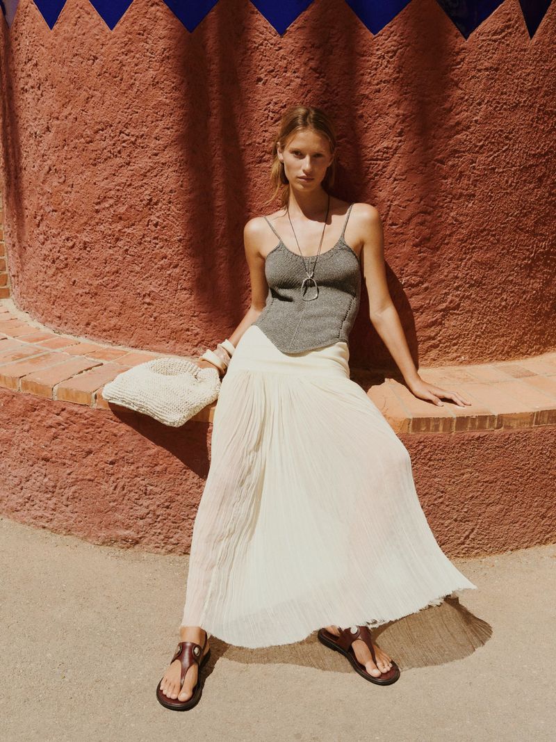 A woman sits wearing a gray tank top, a white skirt, and brown sandals, while posing against a textured, terracotta-colored wall outdoors.