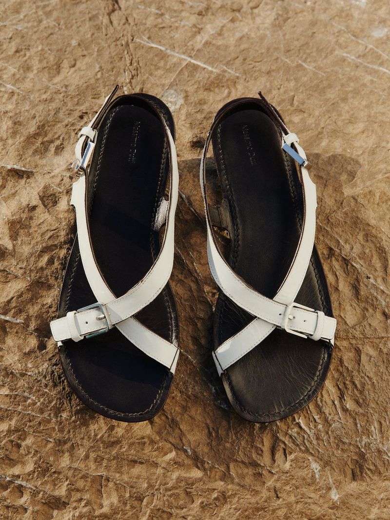 Elevated view of two black leather sandals with white straps and silver buckles, lying on a textured brown rock surface.