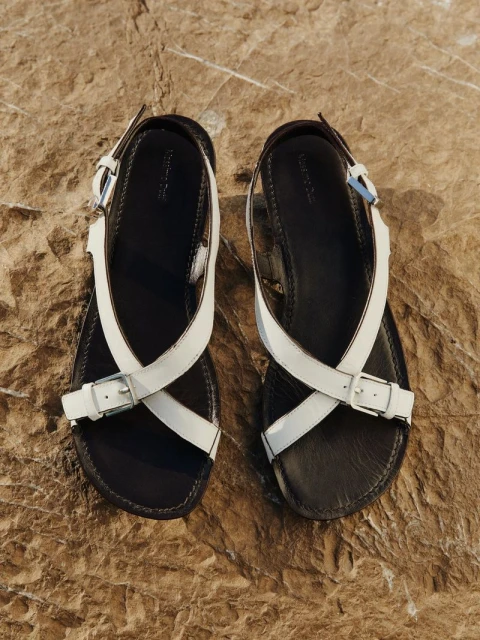 Elevated view of two black leather sandals with white straps and silver buckles, lying on a textured brown rock surface.