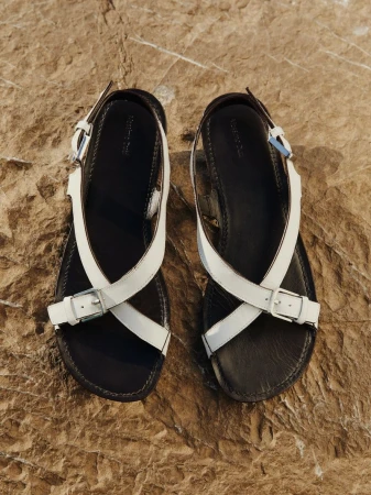 Elevated view of two black leather sandals with white straps and silver buckles, lying on a textured brown rock surface.