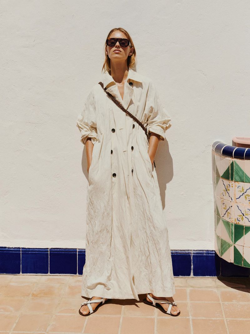 A blonde woman in sunglasses and a light-colored coat poses on a tiled surface in bright daylight, casting a faint shadow on the wall.