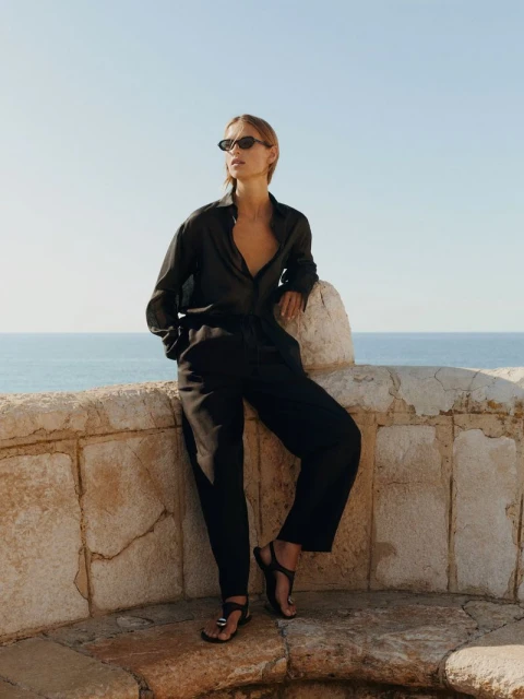 Woman in a black linen outfit and sunglasses posing on stone wall with ocean background, illuminated by natural light in outdoor setting.