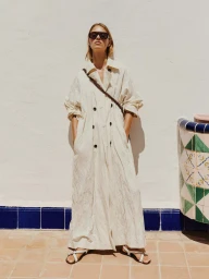 Lea Seelenmeyer wearing an oversized floor-length cream coat, dark sunglasses, and flat sandals against a sunlit white wall.
