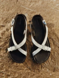 A pair of white leather criss-cross strap sandals with silver buckles resting on a textured brown rock surface.