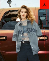Woman with curly blonde hair wearing a grey button-down shirt and dark pants standing in front of a red vehicle
