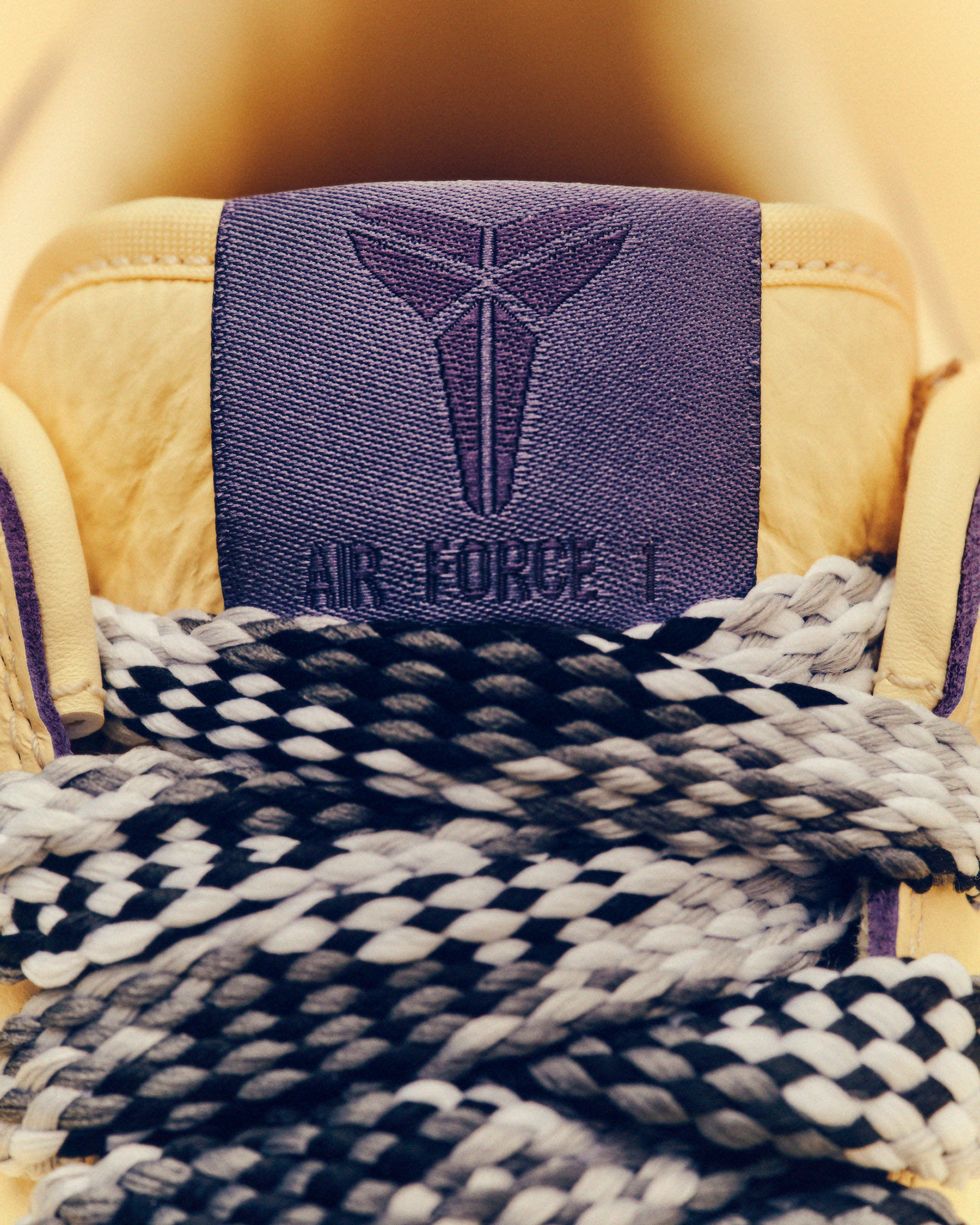 Close-up of a tan Nike Air Force 1 sneaker with black and white laces and a purple tongue featuring the Kobe Bryant logo.