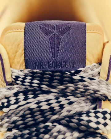 Close-up of a tan Nike Air Force 1 sneaker with black and white laces and a purple tongue featuring the Kobe Bryant logo.