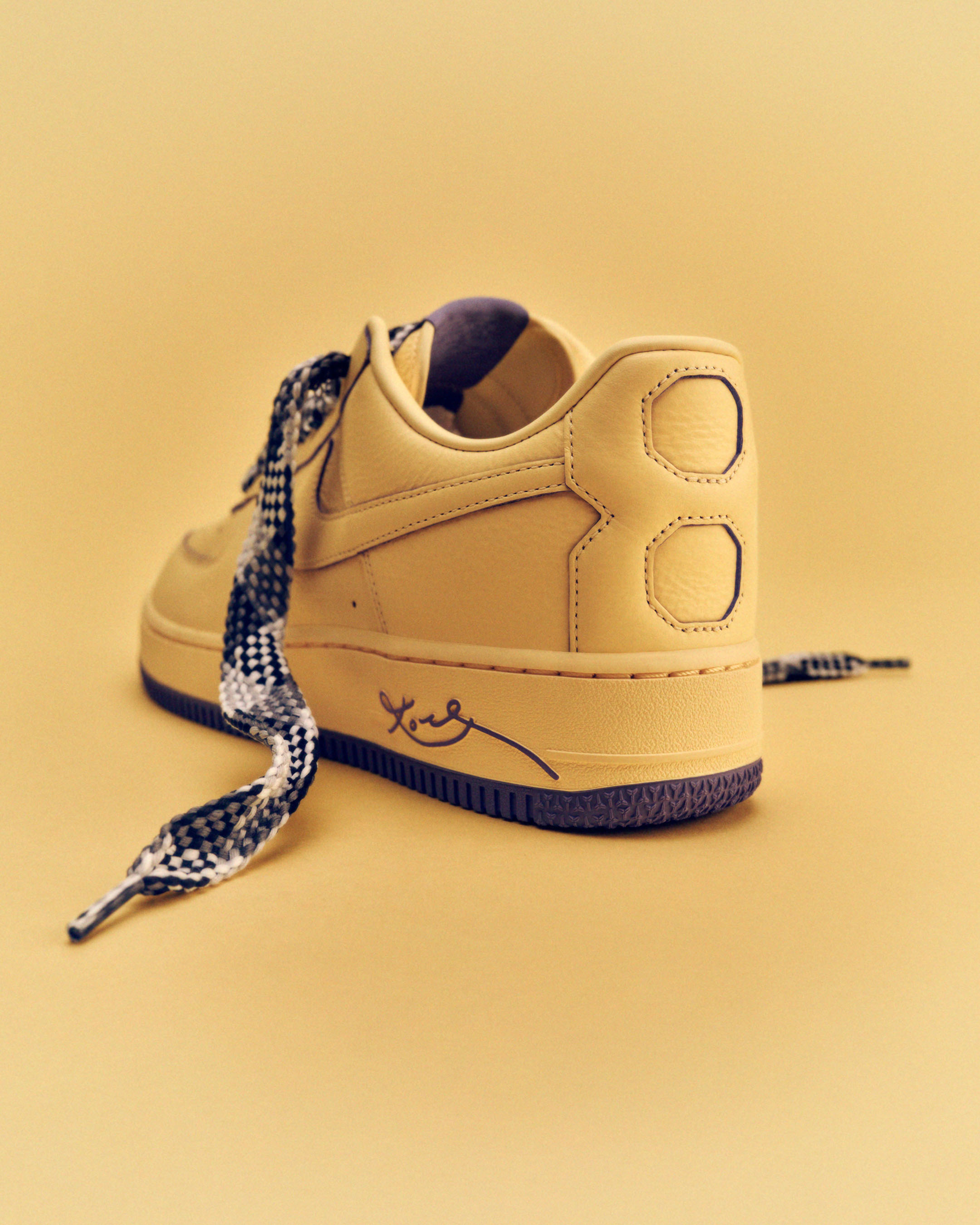 Close-up of a golden-tan Nike Air Force 1 sneaker, showcasing patterned laces and a handwritten-style logo on a matching monochromatic background.