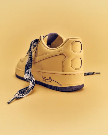 Close-up of a golden-tan Nike Air Force 1 sneaker, showcasing patterned laces and a handwritten-style logo on a matching monochromatic background.