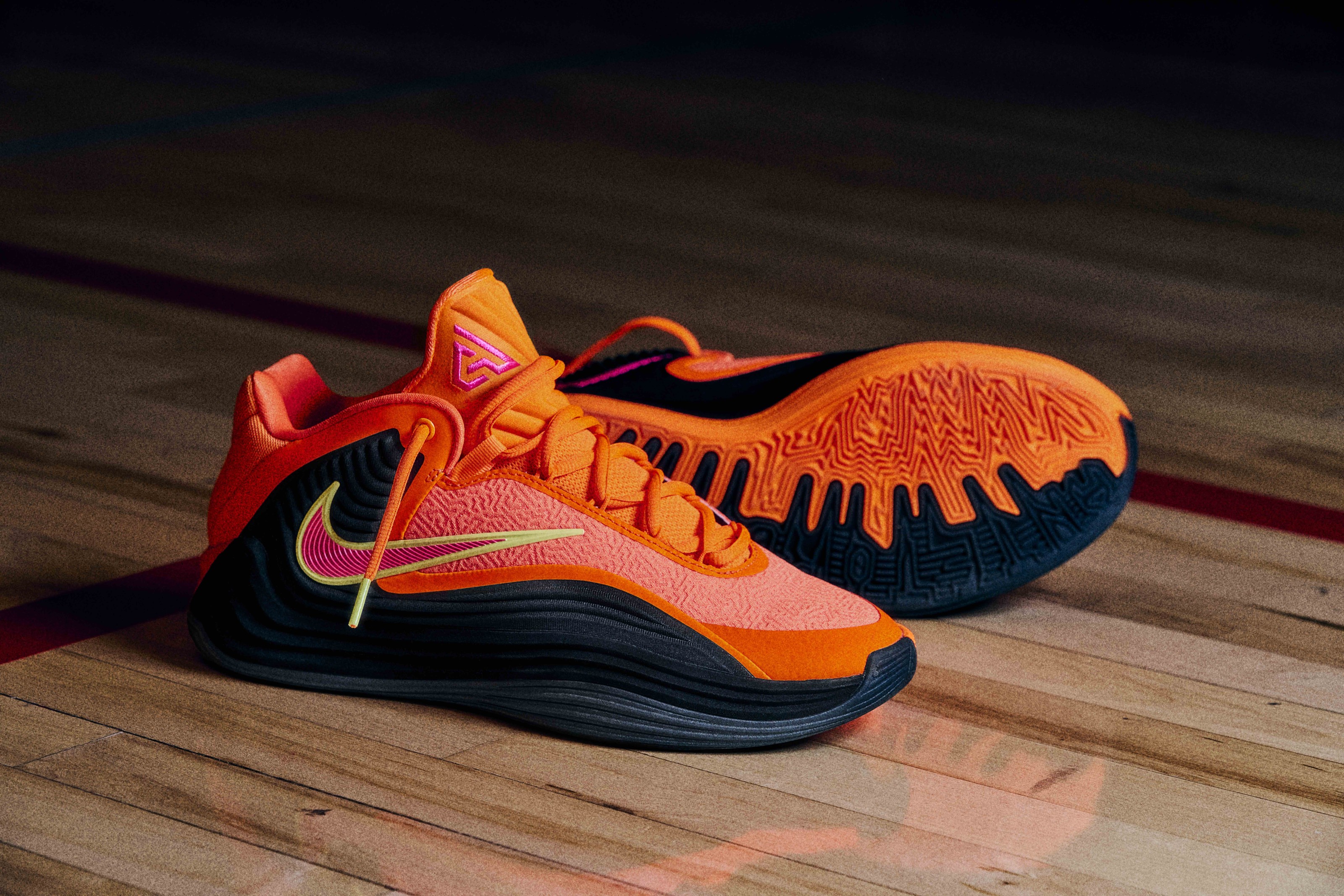 Close-up of an orange and black basketball shoe with pink and yellow accents on a wooden basketball court surface.