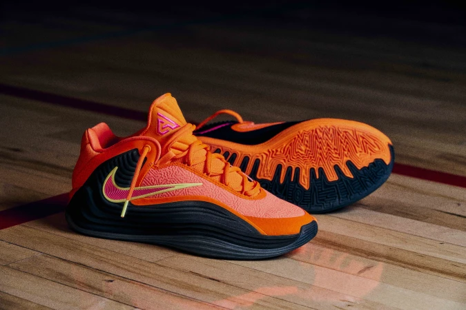 Close-up of an orange and black basketball shoe with pink and yellow accents on a wooden basketball court surface.