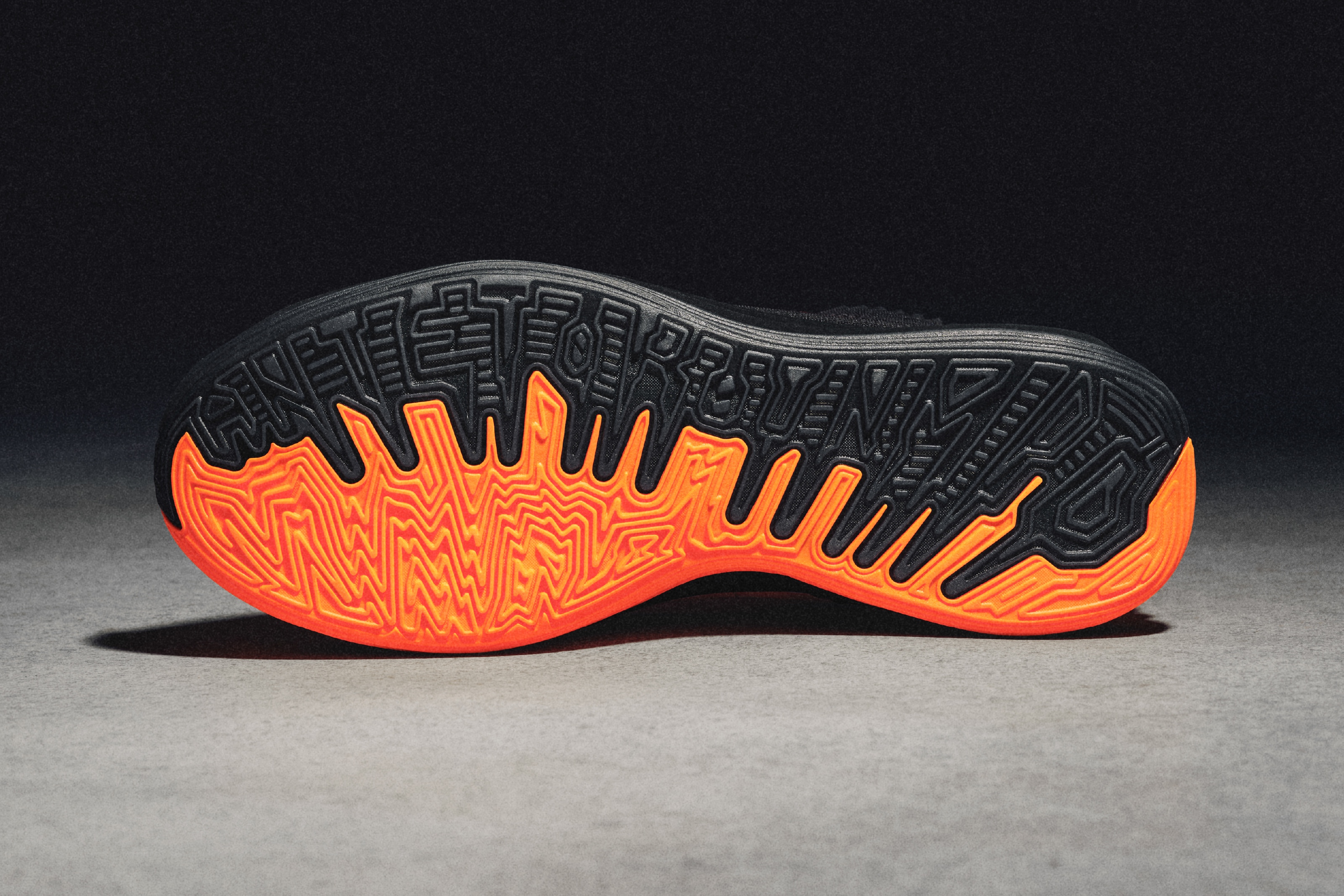 A close-up studio shot showcases the sole of a shoe with intricate geometric patterns in orange and black, positioned against a gray surface.