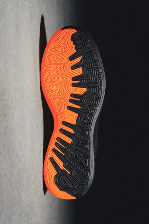 A close-up studio shot showcases the sole of a shoe with intricate geometric patterns in orange and black, positioned against a gray surface.