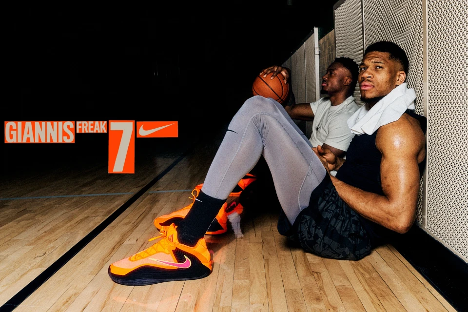Two male athletes sitting on a basketball court with orange athletic shoes and basketball. Nike and Giannis Freak branding.