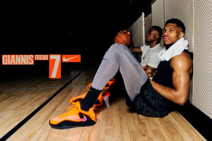Two male athletes sitting on a basketball court with orange athletic shoes and basketball. Nike and Giannis Freak branding.