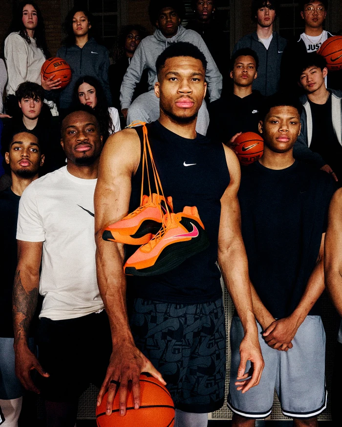 Giannis Antetokounmpo holding a basketball with orange sneakers draped over his shoulder, surrounded by a group of players.