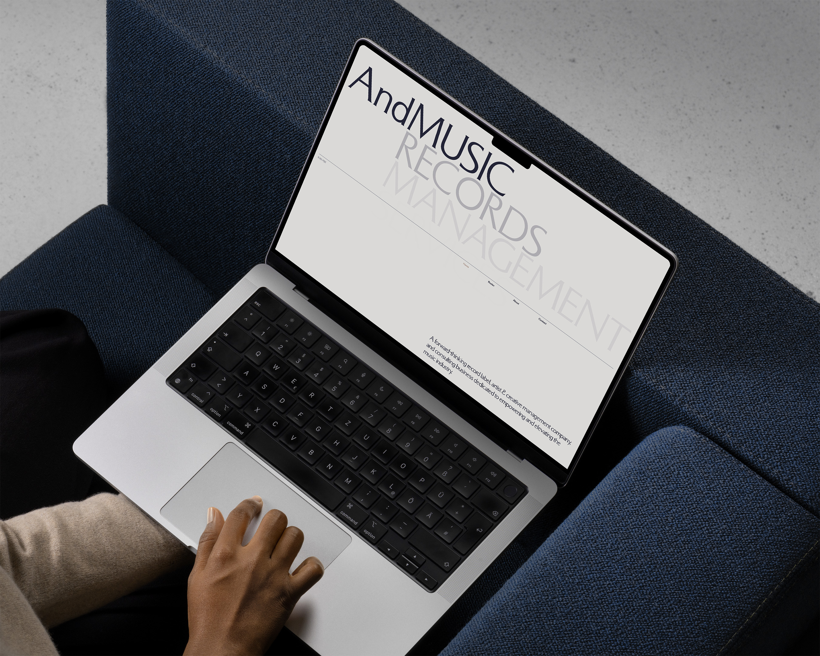 Person using a laptop with 'AndMUSIC Records Management' displayed on screen, set on a blue couch with a hand on the trackpad.