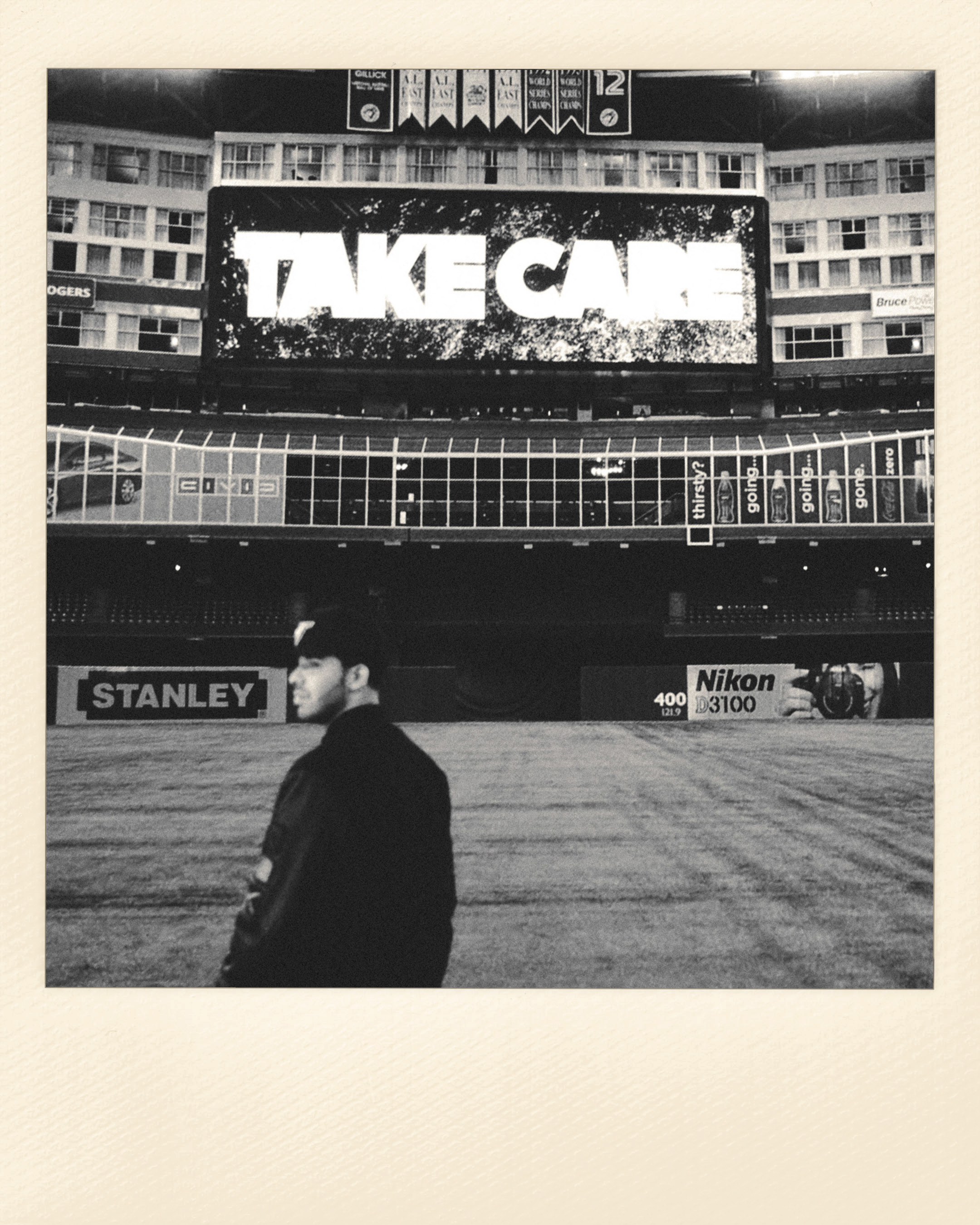 Black and white shot of a man in a baseball cap and jacket standing on a baseball field with a lit-up stadium in the background.