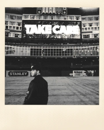 Black and white shot of a man in a baseball cap and jacket standing on a baseball field with a lit-up stadium in the background.