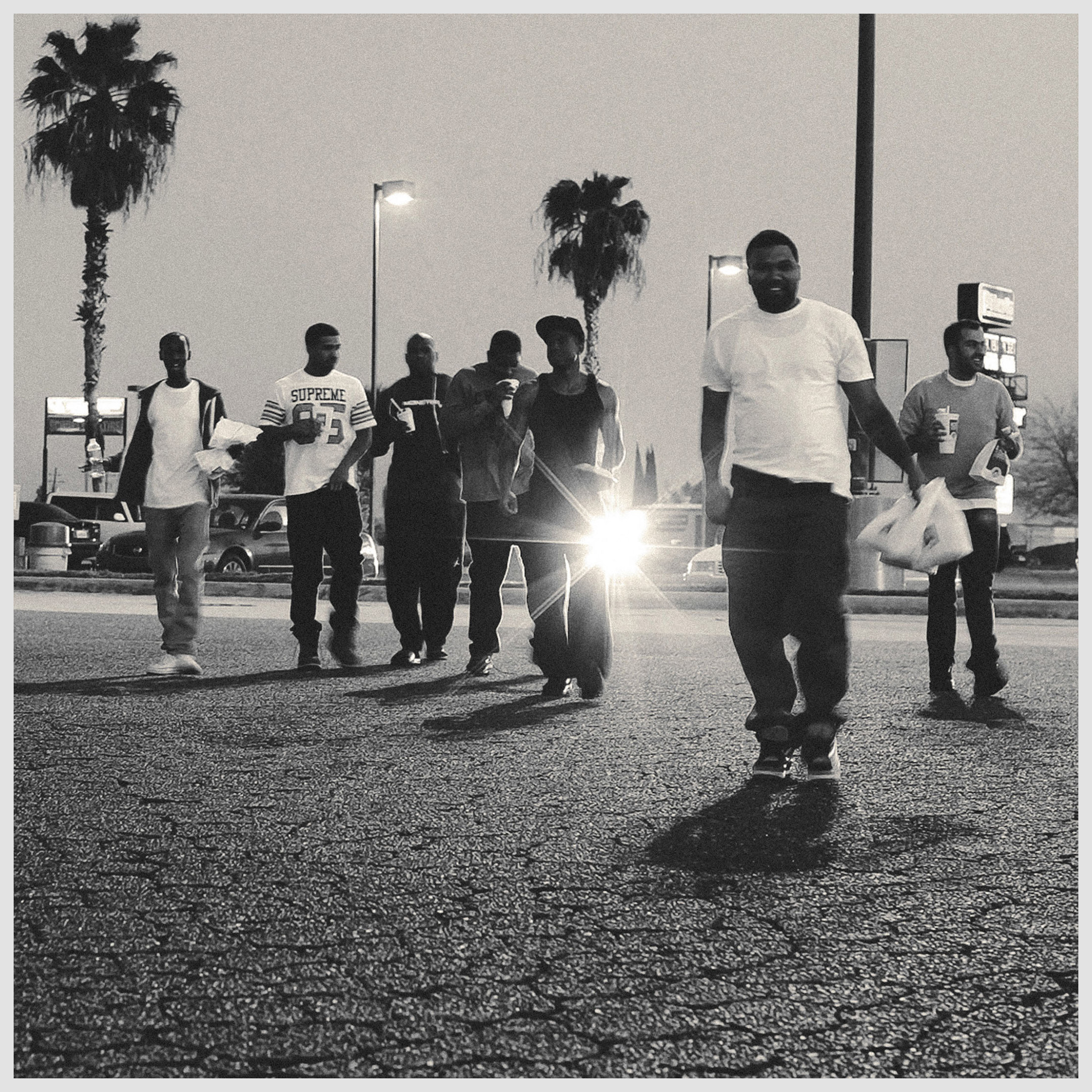 A group of seven men are walking on an asphalt road in a black and white image; several palm trees are visible, as well as cars in the background.
