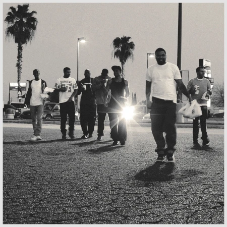 A group of seven men are walking on an asphalt road in a black and white image; several palm trees are visible, as well as cars in the background.
